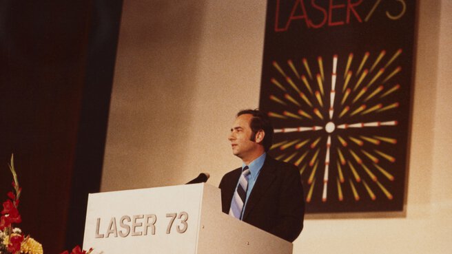 Man in suit and striped tie speaks at a podium. A sign with the 