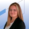 Eine junge Frau mit langen blonden Haaren, Brille und einem schwarzen Blazer lächelt vor einem blauen geometrischen Hintergrund.