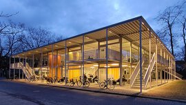 Modernes zweistöckiges Gebäude mit Glaswänden, draußen geparkten Fahrrädern und warmer Beleuchtung vor dem Hintergrund eines Abendhimmels.