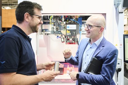 Zwei Männer unterhalten sich freundlich vor einer industriellen Lasermaschine; einer hält ein Metallbauteil in der Hand, während der andere mit Tablet und Anzug technische Details erklärt.