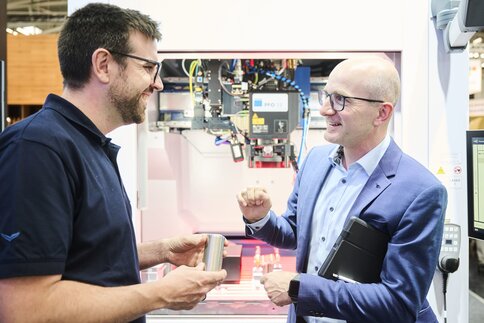 Zwei Männer unterhalten sich freundlich vor einer industriellen Lasermaschine; einer hält ein Metallbauteil in der Hand, während der andere mit Tablet und Anzug technische Details erklärt.