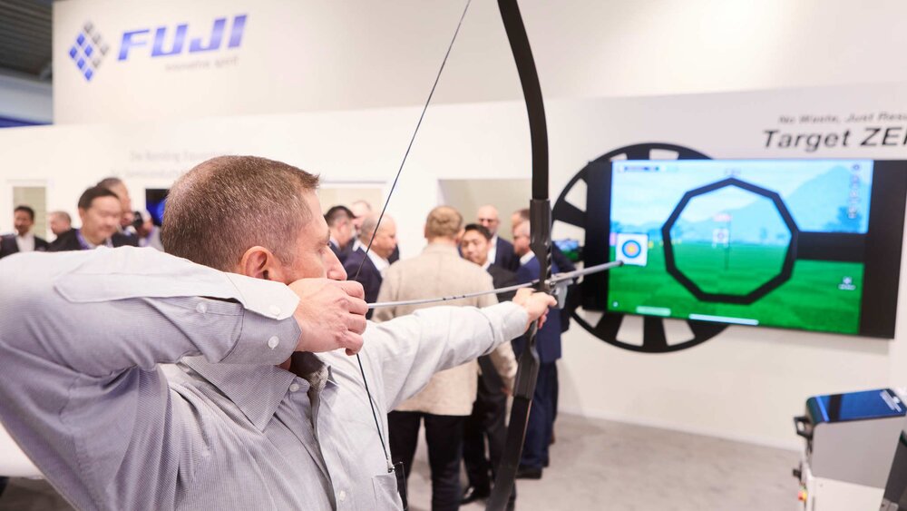 Ein Mann zielt mit Pfeil und Bogen auf einen Bildschirm an einem Messestand, der mehrere Ziele darstellt. Im Hintergrund sind weitere Besucher zu sehen.