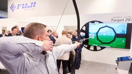 Ein Mann zielt mit Pfeil und Bogen auf einen Bildschirm an einem Messestand, der mehrere Ziele darstellt. Im Hintergrund sind weitere Besucher zu sehen.