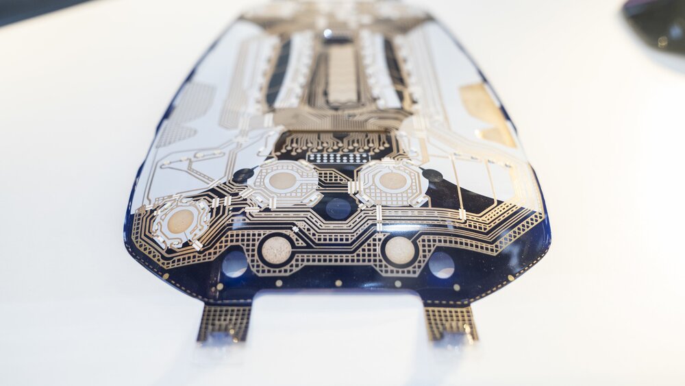 A close-up view of a printed circuit board in black, white and beige with rounded edges lies on a white background.