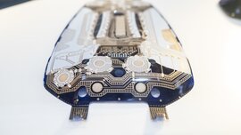 A close-up view of a printed circuit board in black, white and beige with rounded edges lies on a white background.