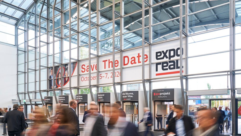 Eine Menschenmenge schreitet durch ein modernes Kongresszentrum aus Glas; über den Eingangsständen hängen große Banner mit der Aufschrift „Save The Date: 5.–7. Oktober 2022 und ‚Expo Real‘“.