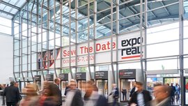 Eine Menschenmenge schreitet durch ein modernes Kongresszentrum aus Glas; über den Eingangsständen hängen große Banner mit der Aufschrift „Save The Date: 5.–7. Oktober 2022 und ‚Expo Real‘“.