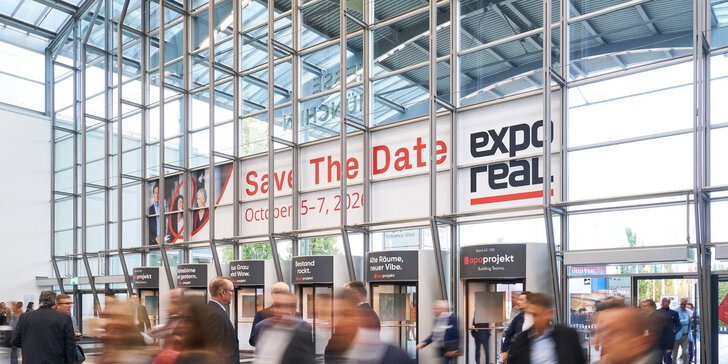 Eine Menschenmenge schreitet durch ein modernes Kongresszentrum aus Glas; über den Eingangsständen hängen große Banner mit der Aufschrift „Save The Date: 5.–7. Oktober 2022 und ‚Expo Real‘“.