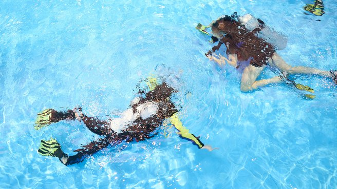 View from above of an indoor pool in which two people in diving gear are diving underwater.