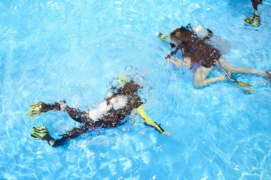View from above of an indoor pool in which two people in diving gear are diving underwater.