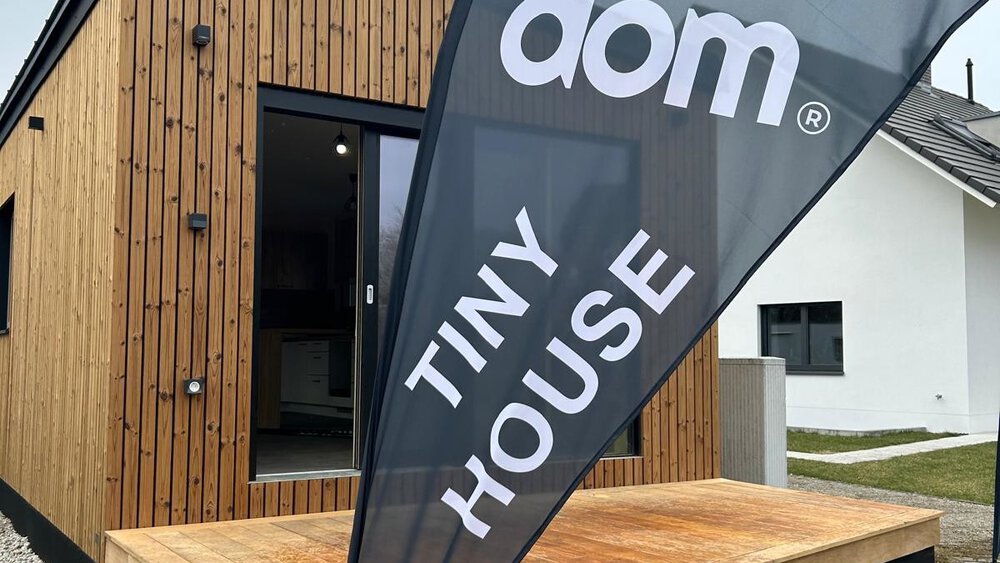 Ein schmales Tiny House aus Holz vor dem ein schwarzer Werbebanner mit den Worten „Kolodom“ und „Tiny House“ steht.