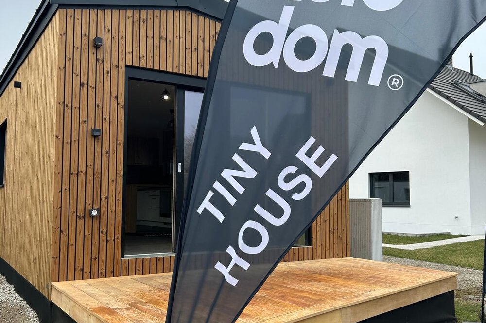 Ein schmales Tiny House aus Holz vor dem ein schwarzer Werbebanner mit den Worten „Kolodom“ und „Tiny House“ steht.