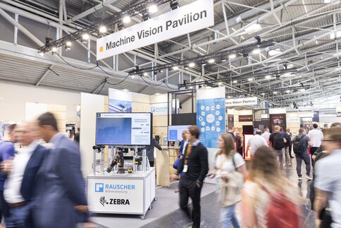 Besucher, die über die Ausstellung Machine Vision Pavilion laufen. Im Hintergrund sind Mesestände zu sehen