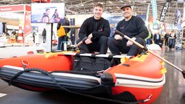 Two smiling men dressed in black are sitting in a red rubber dinghy at an indoor trade fair or exhibition.