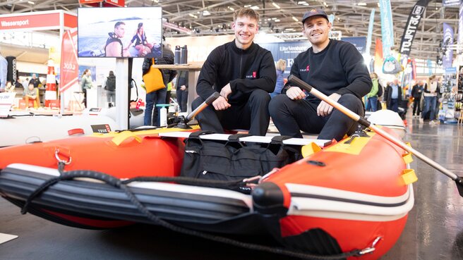 Zwei lächelnde Männer in schwarzer Kleidung sitzen in einem roten Schlauchboot auf einer Indoor-Messe oder einer Fachmesse.