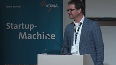 Ein Mann mit Brille und Blazer trägt an einem Podium vor einem 