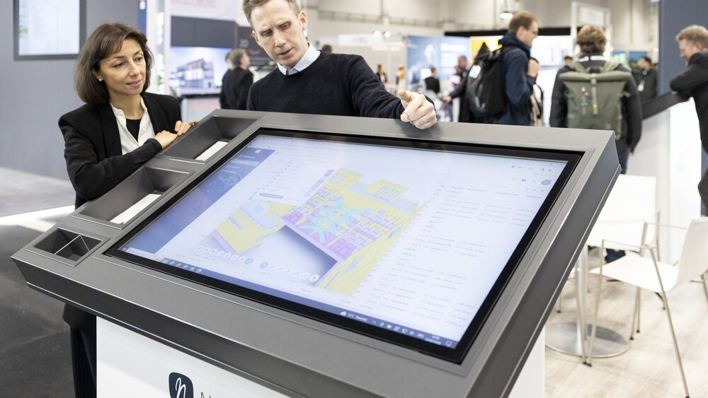 Zwei Personen diskutieren über einen digitalen Grundriss, der auf einem großen Touchscreen an einem technischen Messestand angezeigt wird.