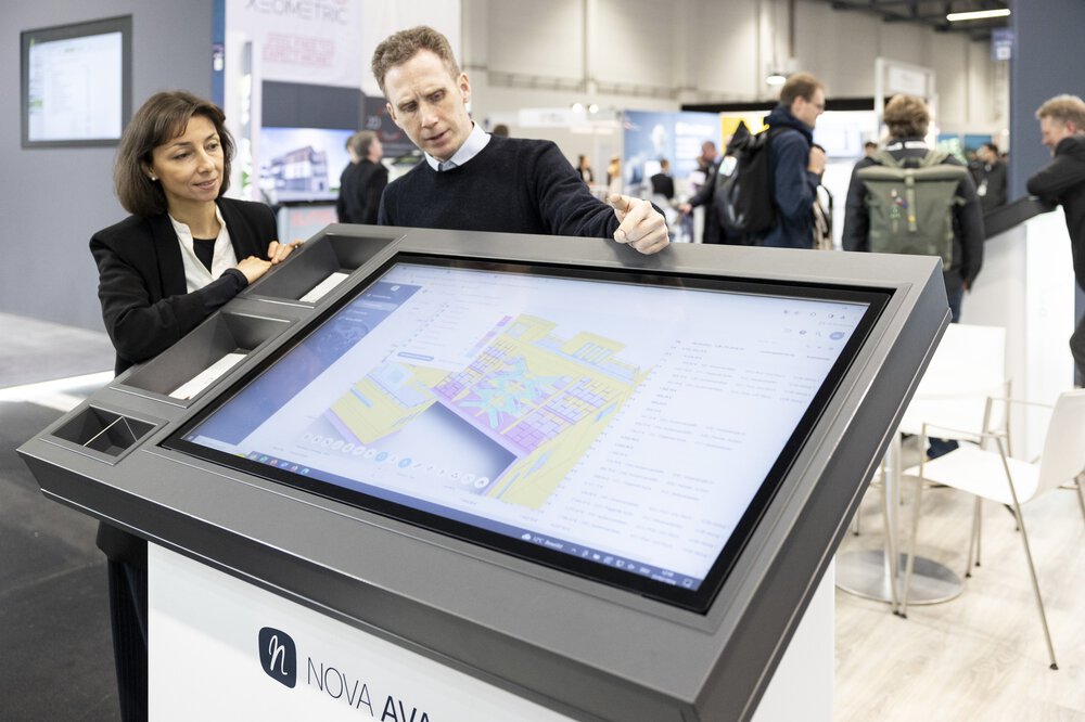 Zwei Personen diskutieren über einen digitalen Grundriss, der auf einem großen Touchscreen an einem technischen Messestand angezeigt wird.