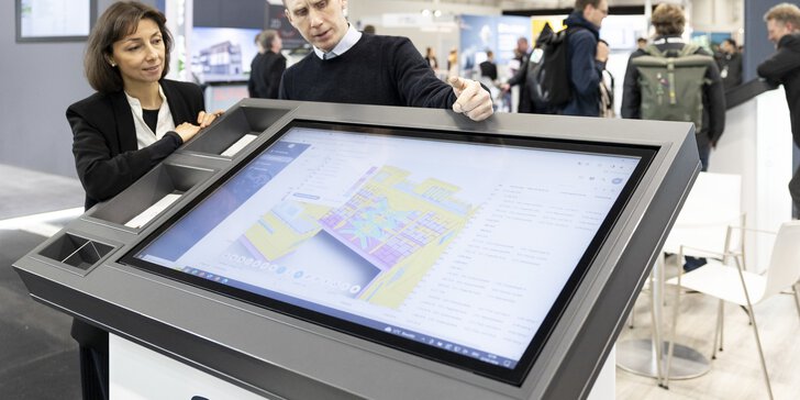 Zwei Personen diskutieren über einen digitalen Grundriss, der auf einem großen Touchscreen an einem technischen Messestand angezeigt wird.