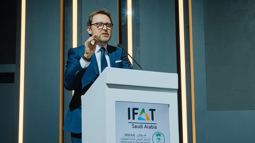  A man wearing glasses and a blue suit stands at a lectern at IFAT Saudi Arabia and speaks to an audience outside the frame.
