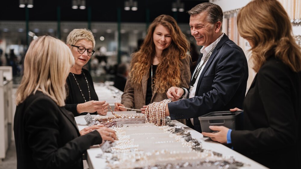 A man and four women are standing around a display table, looking at various pearl necklaces. The man is holding up a few necklaces with a smile.