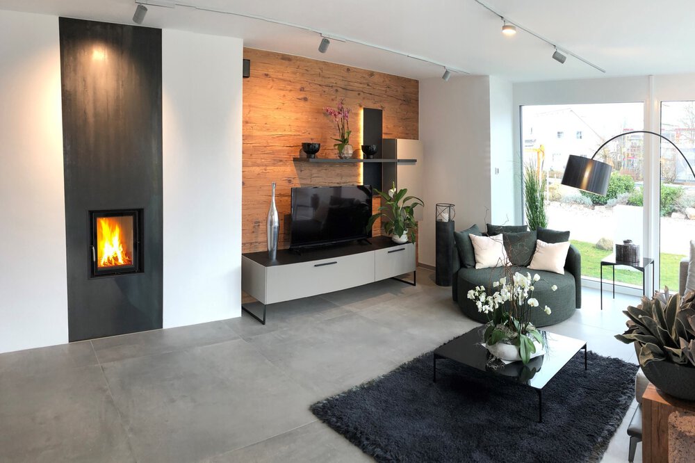 Modernes Wohnzimmer mit Kamin, Fernseher, grauem Sofa, schwarzem Teppich, Pflanzen und großen Fenstern mit Blick auf einen Garten.