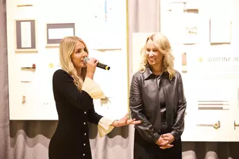 Two women speak in front of a framed artwork on a wall, holding a microphone and smiling at an indoor event.