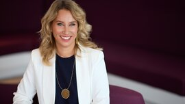 Eine lächelnde Frau mit langen gewellten blonden Haaren trägt Ohrringe und eine Halskette, eine blaue Bluse und einen weißen Blazer.