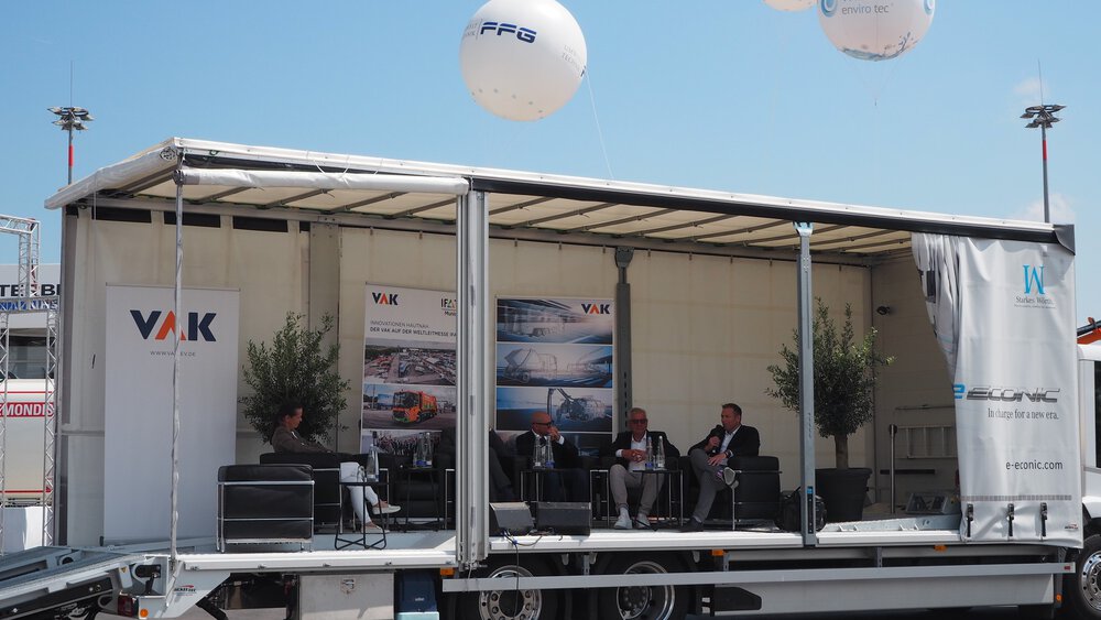 Podiumsdiskussion auf der IFAT Munich in einem offenen Trailer-Messestand von VAK und Partnern, mit Bannern, Sitzmöbeln und Branding unter freiem Himmel.