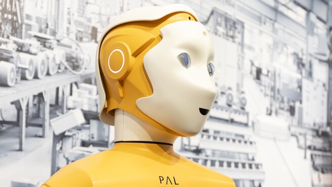 Ein gelber humanoider Roboter mit einem Touchscreen auf der Brust vor einem Foto einer Fabrikhalle.
