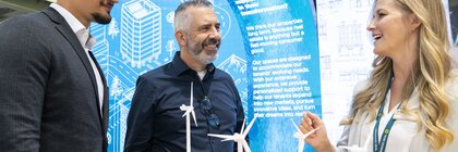 Three people talking at a trade fair stand, in front of a stepped wooden model on which several wind turbines stand.