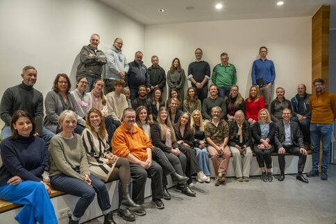Zahlreiche Personen sitzen und stehen in drei Reihen für ein Gruppenfoto in einem Raum mit beigen Wänden und lächeln.