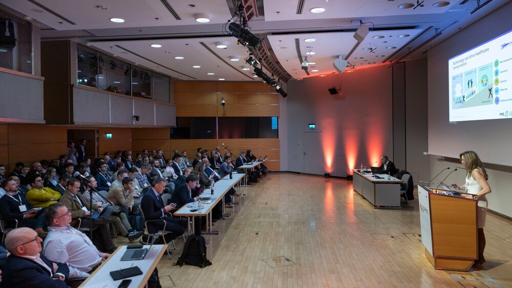 A well-attended conference hall with a man at a laptop and a woman at a lectern giving a presentation to an audience.