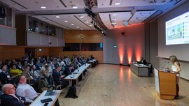 A well-attended conference hall with a man at a laptop and a woman at a lectern giving a presentation to an audience.