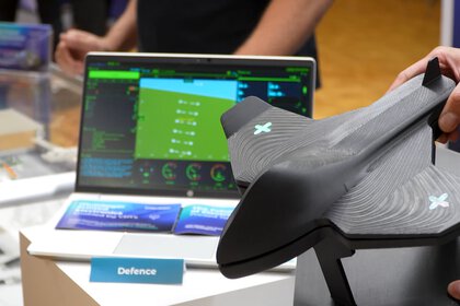 Eine Nahaufnahme auf der Technologiemesse LOPEC. Im Vordergrund hält eine Person ein schwarzes, aerodynamisches 3D-Modell eines Flugobjekts (Drohne oder Jet) mit leuchtenden X-Symbolen auf den Tragflächen. Im Hintergrund ist ein Laptop mit einer grün-blauen Benutzeroberfläche und Daten-Dashboards sowie ein kleines Schild mit der Aufschrift „Defence“ zu sehen.