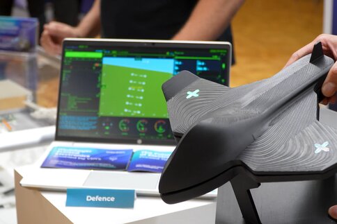 Eine Nahaufnahme auf der Technologiemesse LOPEC. Im Vordergrund hält eine Person ein schwarzes, aerodynamisches 3D-Modell eines Flugobjekts (Drohne oder Jet) mit leuchtenden X-Symbolen auf den Tragflächen. Im Hintergrund ist ein Laptop mit einer grün-blauen Benutzeroberfläche und Daten-Dashboards sowie ein kleines Schild mit der Aufschrift „Defence“ zu sehen. 