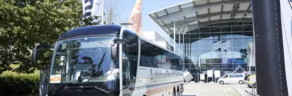 A white coach stands on the cobblestones in front of an entrance building to the Munich Trade Fair Centre. Colourful flags are flying next to it.