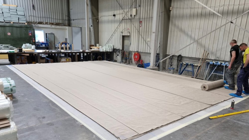 Zwei Männer rollen eine große beige Plane auf dem Boden einer geräumigen Industriehalle aus