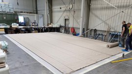 Zwei Männer rollen eine große beige Plane auf dem Boden einer geräumigen Industriehalle aus