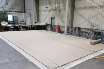 Two men unroll a large beige tarpaulin on the floor of a spacious industrial hall