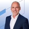 Lächelnder Mann in Anzug und weißem Hemd, der vor einem blauen geometrischen Hintergrund steht.