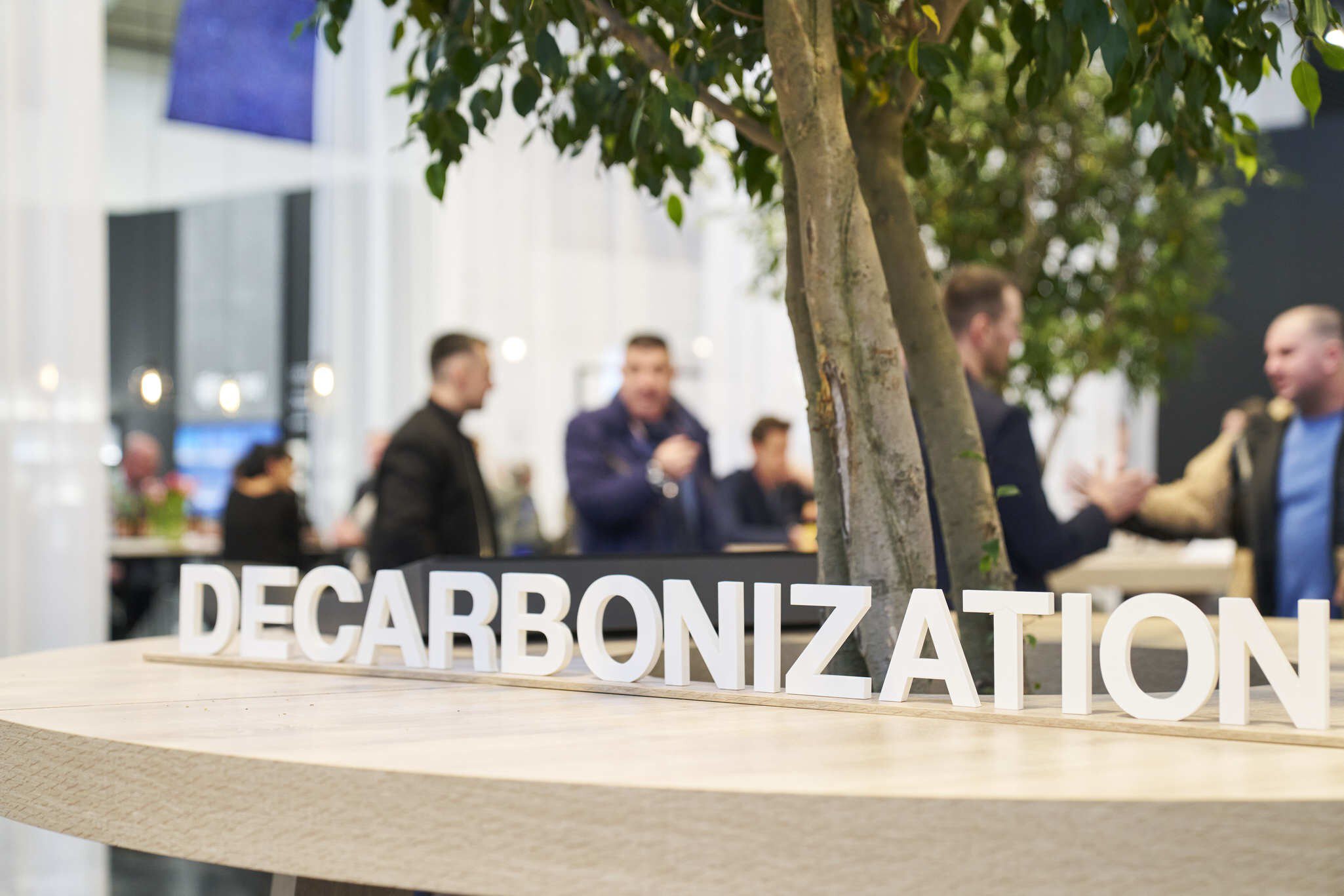 The word ‘DECARBONIZATION’ is written in white letters on a wooden table from which a small tree is growing. Visitors to the trade fair can be seen in the background.