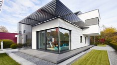 Modernes weißes Haus mit großen Fenstern, Flachdach und Holzterrasse, umgeben von einem gepflegten Rasen und einem Kiesweg.