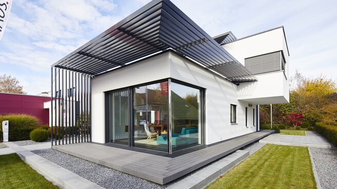 Modernes weißes Haus mit großen Fenstern, Flachdach und Holzterrasse, umgeben von einem gepflegten Rasen und einem Kiesweg.