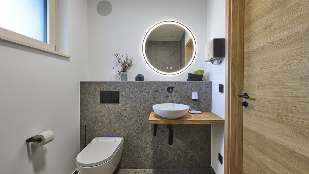 Modernes Badezimmer mit wandmontiertem WC, rundem Spiegel mit LED-Beleuchtung, Aufsatzwaschbecken, Holzakzenten und Steinfliesen.
