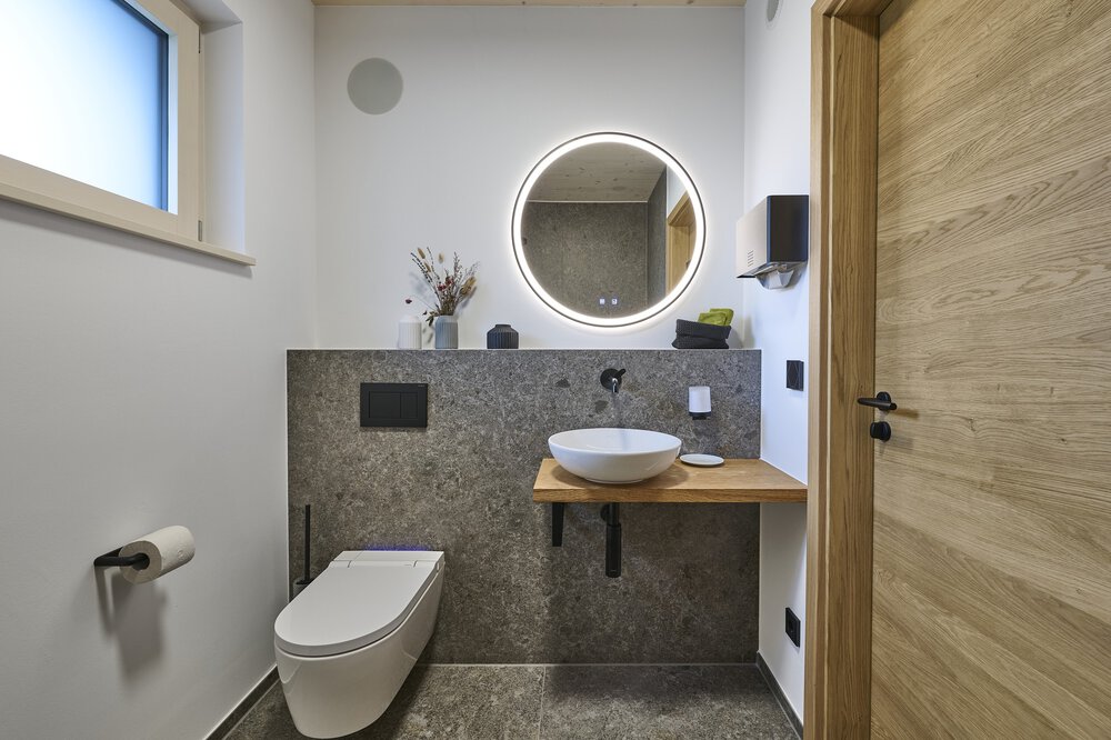 Modernes Badezimmer mit wandmontiertem WC, rundem Spiegel mit LED-Beleuchtung, Aufsatzwaschbecken, Holzakzenten und Steinfliesen.
