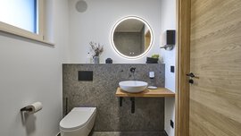 Modernes Badezimmer mit wandmontiertem WC, rundem Spiegel mit LED-Beleuchtung, Aufsatzwaschbecken, Holzakzenten und Steinfliesen.