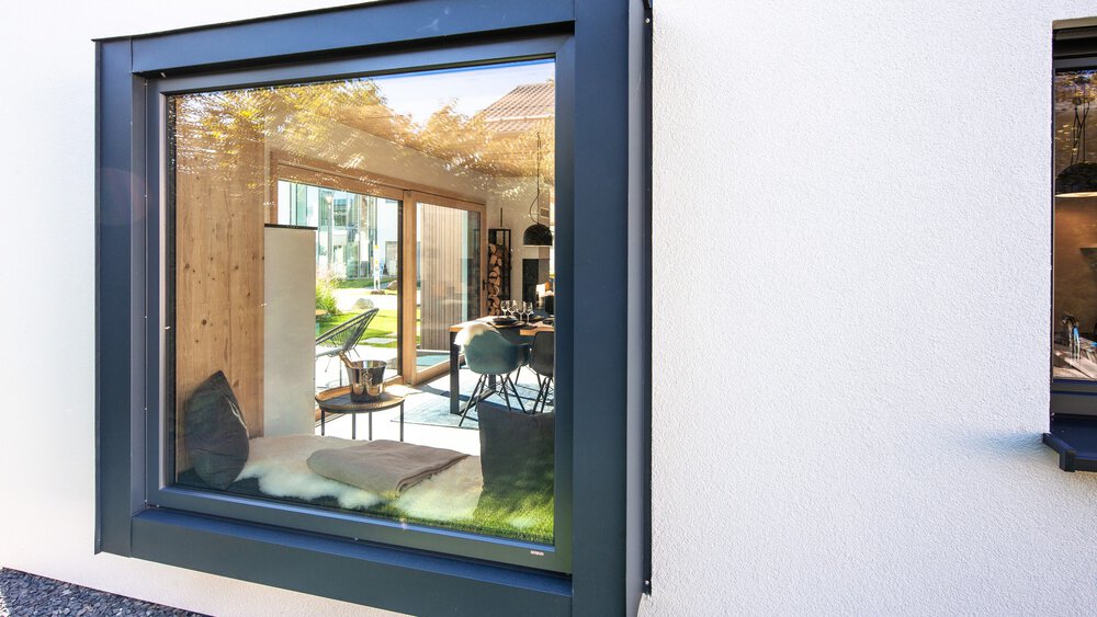 Blick durch ein Fenster auf ein helles Wohnzimmer mit Zimmerpflanzen und einer Terrasse im Freien.