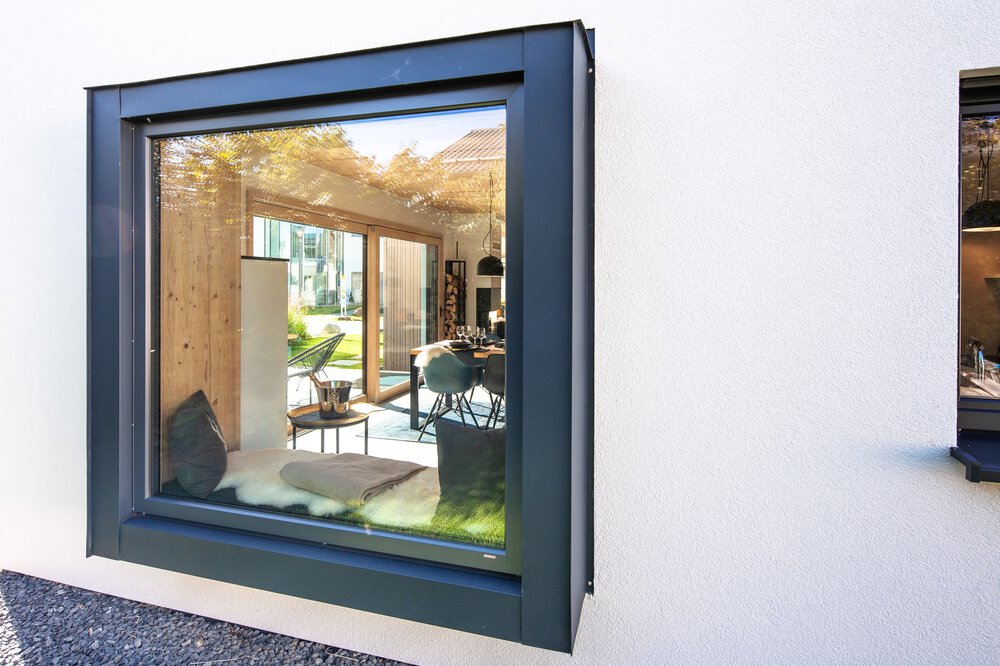 Blick durch ein Fenster auf ein helles Wohnzimmer mit Zimmerpflanzen und einer Terrasse im Freien.