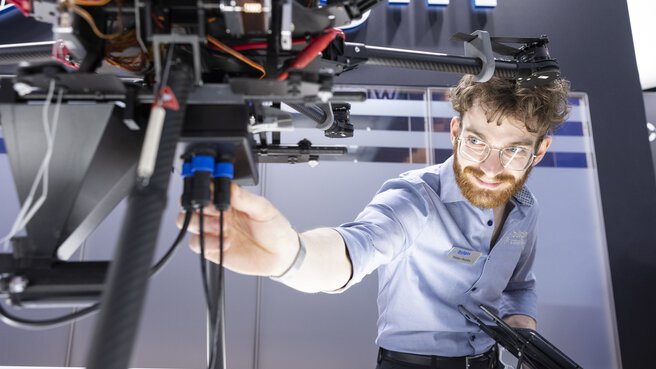 A man with glasses and a light blue shirt connects a cable to a large drone displayed on a pedestal.